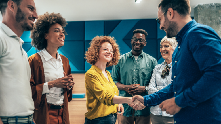 Group of six diverse professionals smiling as one person shakes hands with another, symbolizing teamwork and collaboration.